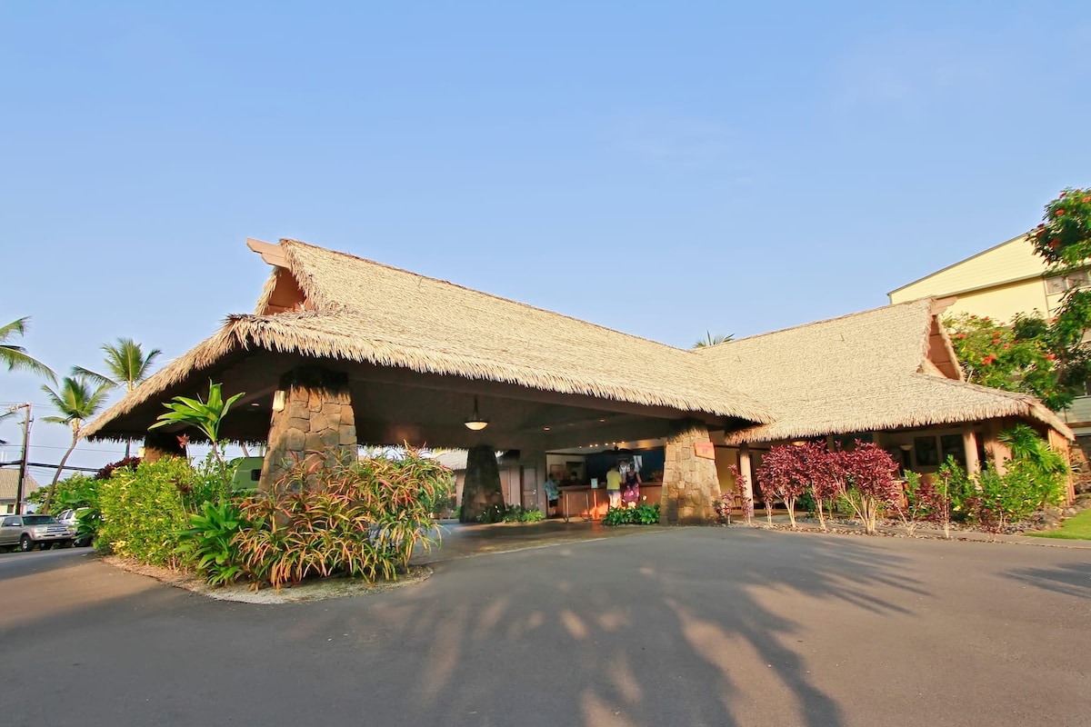 The entrance showcases a traditional Hawaiian hut with a thatched roof, supported by stone columns. Lush tropical plants surround the area, creating a welcoming ambiance. The clear blue sky adds to the overall warmth of the setting, inviting guests to explore the resort.