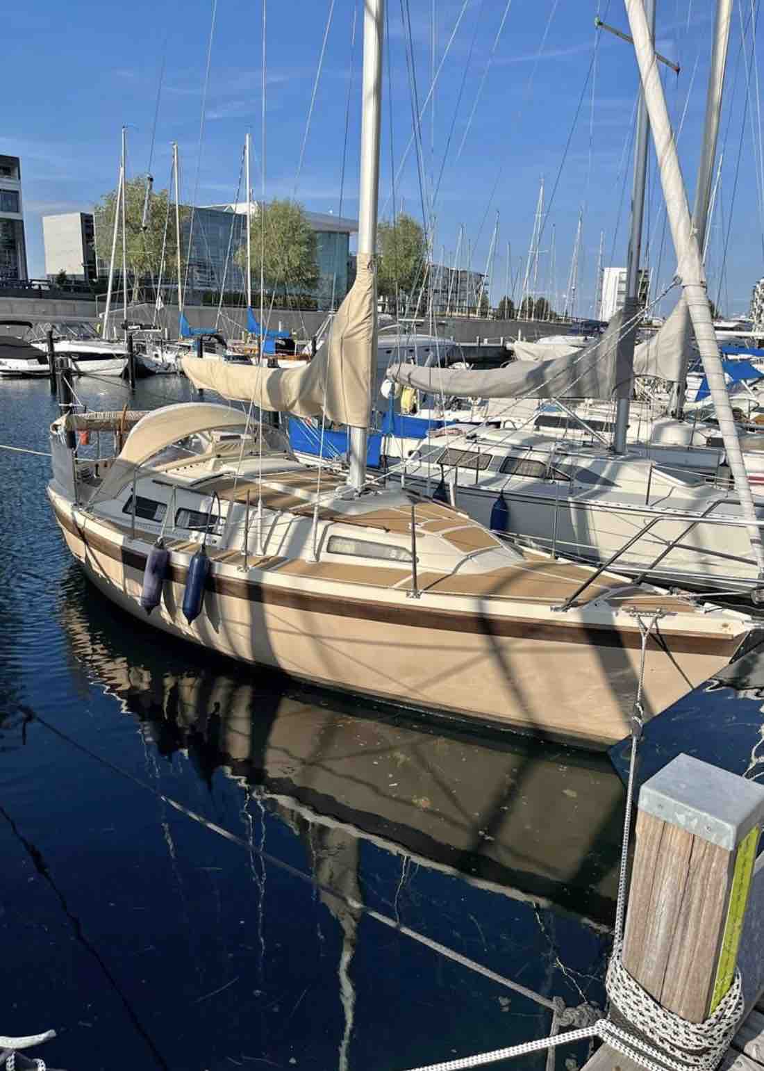 A sleek sailboat is docked in a harbor surrounded by other boats. The wooden exterior is complemented by a soft, mellow finish. Reflections of the boat ripple gently on the water's surface, with masts and sails visible in the background under a clear blue sky.