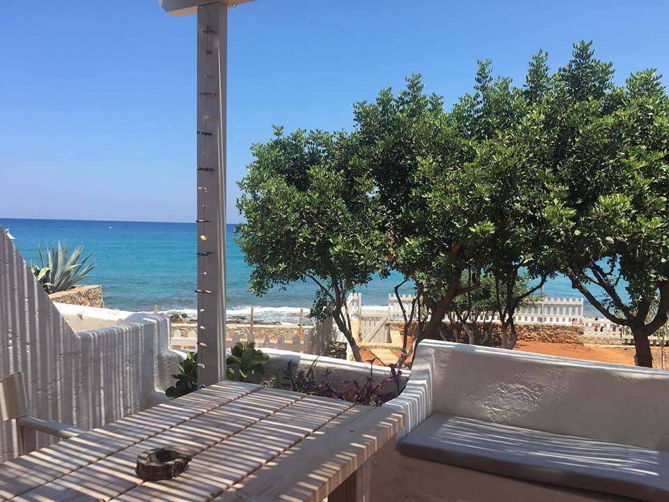 A private outdoor space features a wooden table surrounded by a spacious terrace with white walls. Sea views are framed by greenery, including several trees. The turquoise water is visible just beyond a low boundary wall, creating a serene seaside atmosphere.
