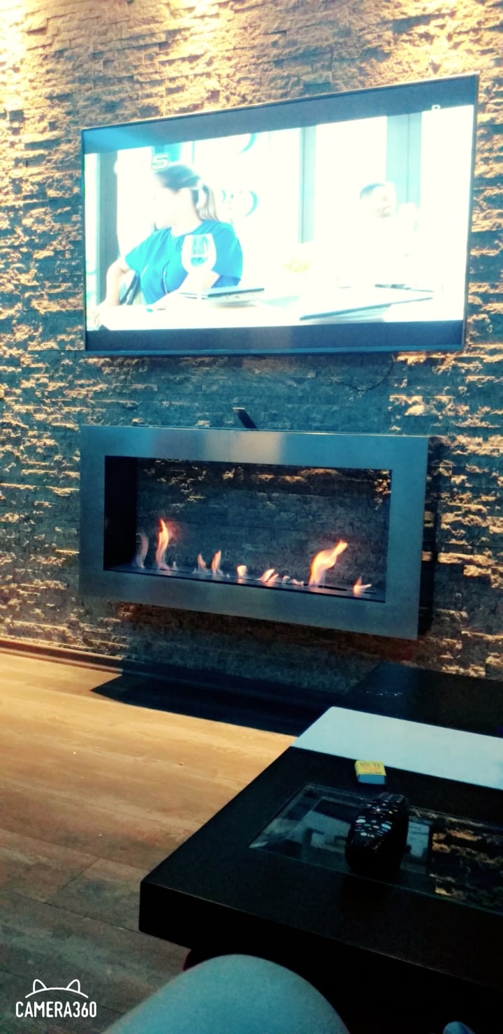 A modern living area is presented with a sleek television mounted above a contemporary fireplace. The stone wall adds texture, while a small coffee table with a remote control is positioned in front. Soft lighting enhances the inviting ambiance of the space.