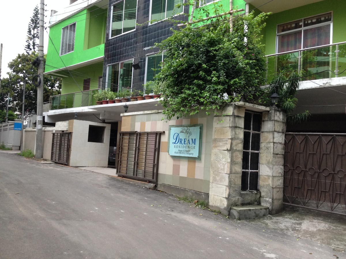The entrance to a residential building is visible, featuring a stone wall with a sign reading 'Dream Residence'. The architectural design exhibits a blend of stone and green elements on the facade. Trees and plants can be seen near the entrance, enhancing the natural surroundings.