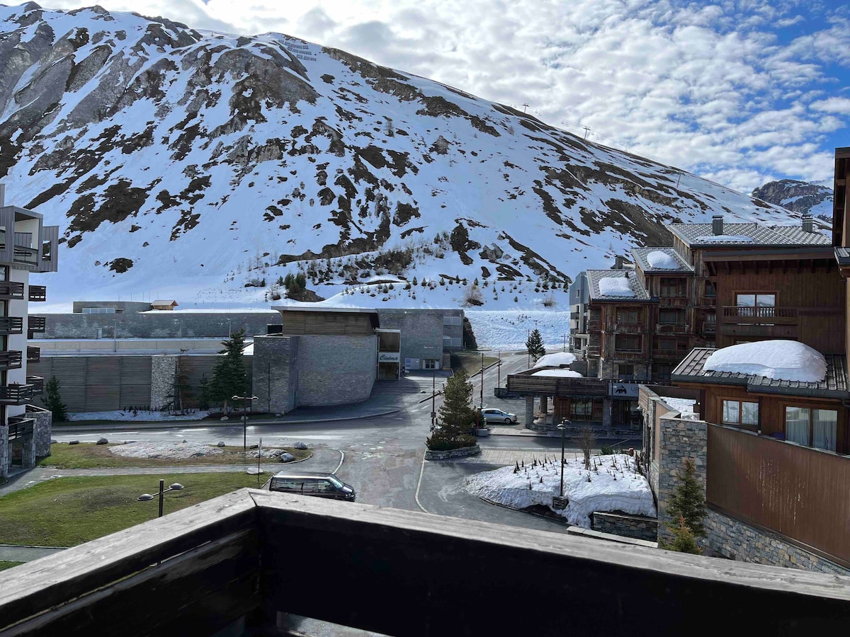 A scenic view captures the snow-covered mountains and resort buildings from a balcony. The foreground features a landscaped area with trees, while the road gently winds through the vibrant alpine setting, creating a serene winter landscape.