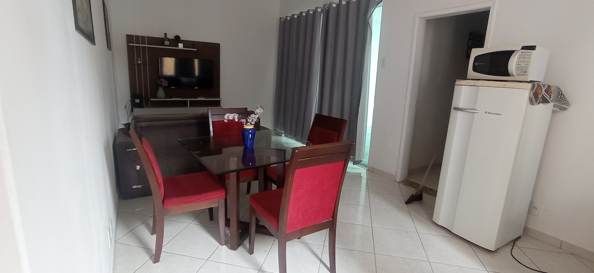 A dining area is provided with a glass-top table surrounded by six red chairs. A wooden sideboard displays a television, and a refrigerator stands in the corner. Natural light enters through a doorway with light-colored curtains, enhancing the simple layout.