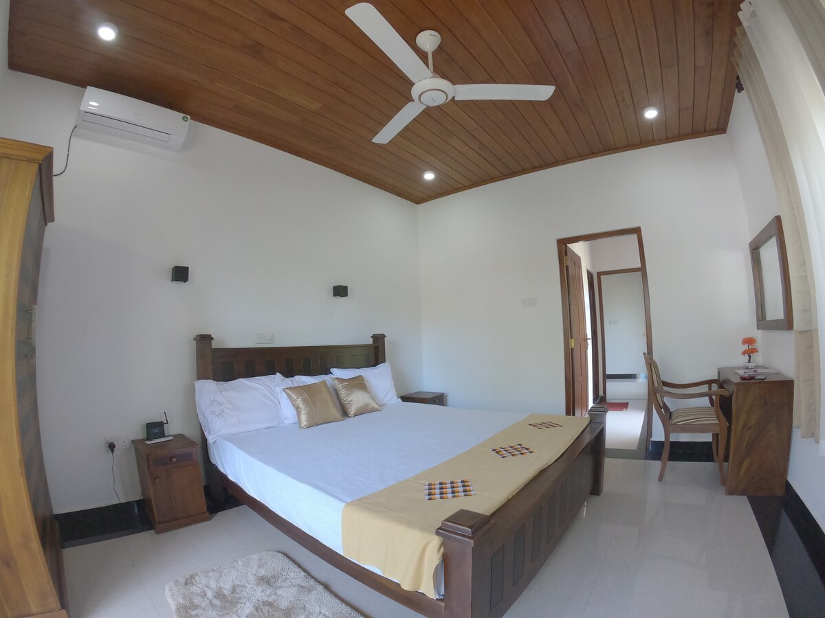 A spacious bedroom is furnished with a large bed featuring neutral tones and decorative pillows. Natural light filters in through a window, and an air conditioning unit is mounted on the wall. A small desk with a chair and a mirror enhances the practical layout of the space.