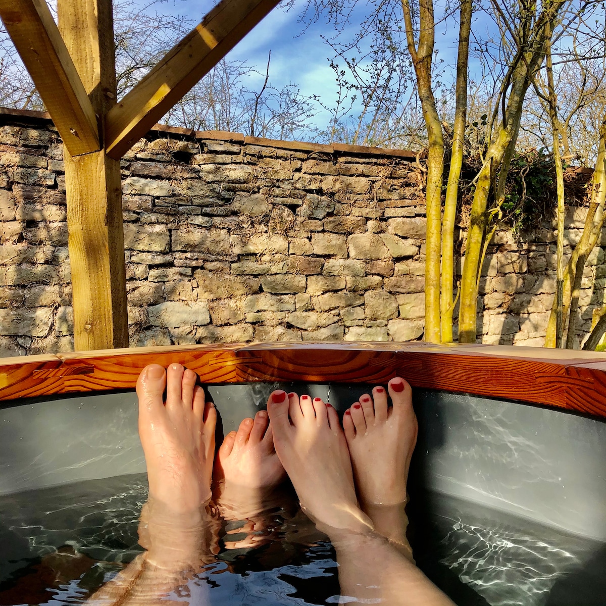 Two pairs of feet are visible in a wood-fired hot tub, with water gently rippling around them. The backdrop of a rustic stone wall and trees under a clear blue sky creates a peaceful outdoor setting. Light plays on the surface of the water, adding to the serene ambiance.