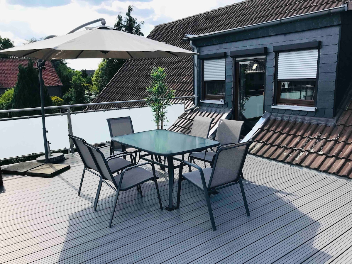A spacious terrace is furnished with a glass-top dining table surrounded by four chairs. An umbrella provides shade, complementing the view of the nearby rooftops. Large windows are visible in the background, offering natural light to the area.