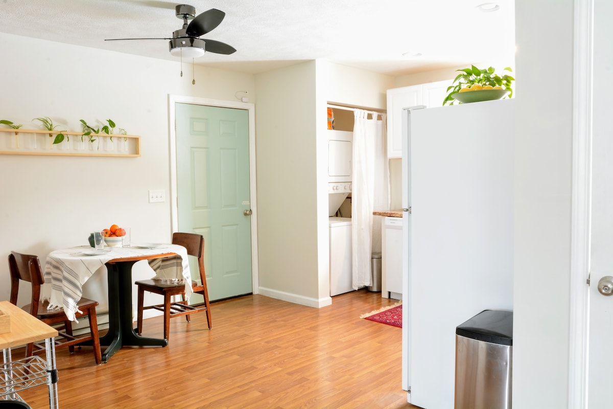 A bright and functional kitchen space features a small dining table with two chairs and a ceiling fan above. A light green door leads to a laundry area, with a white refrigerator and a stainless steel trash bin visible. Natural light illuminates the warm wooden flooring.