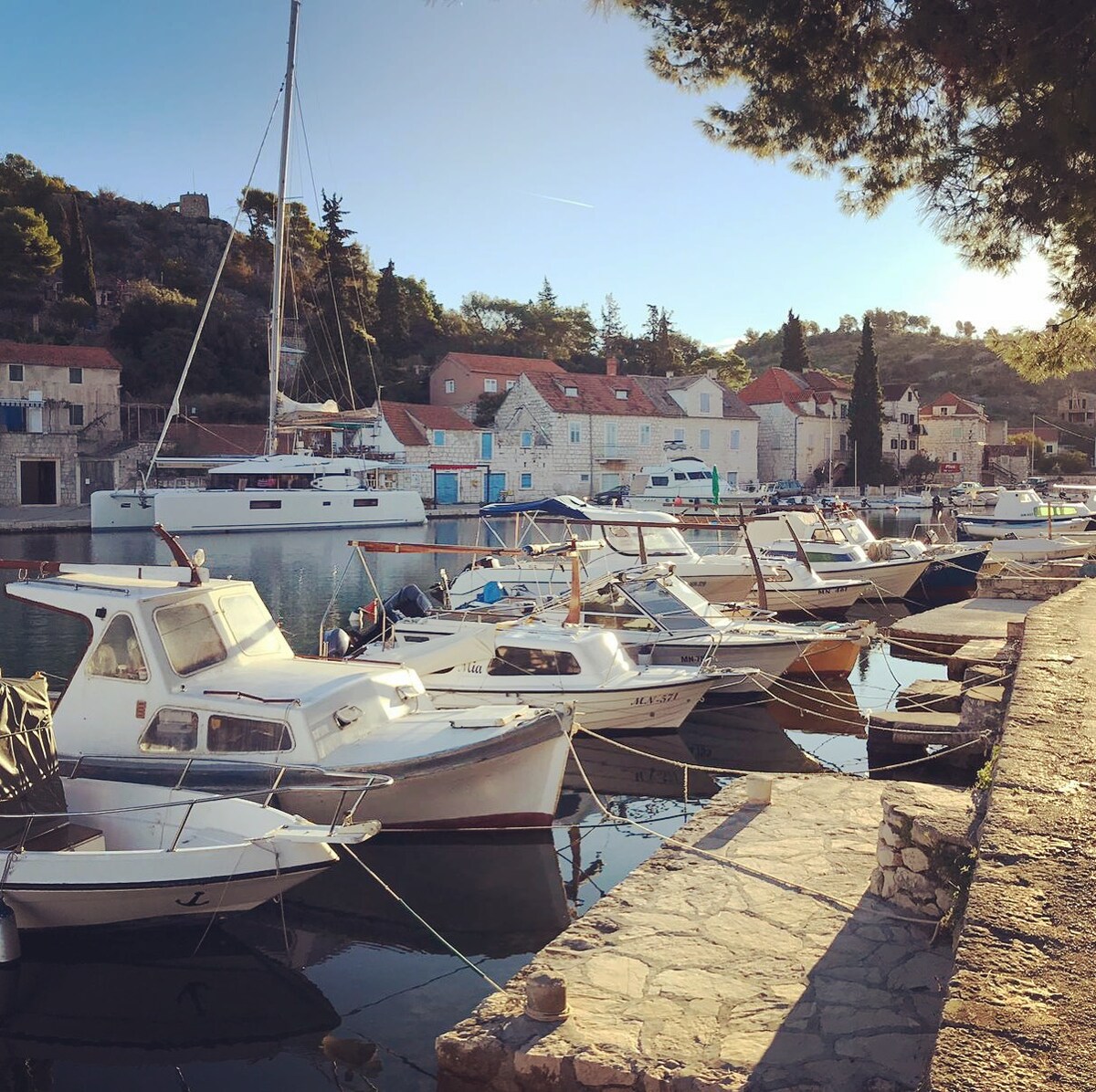 A serene waterfront scene depicts several boats docked along a stone quay. Quaint buildings with tiled roofs emerge in the background, nestled against a wooded hillside. Soft sunlight casts a warm glow on the water, creating reflections from the boats and surroundings.