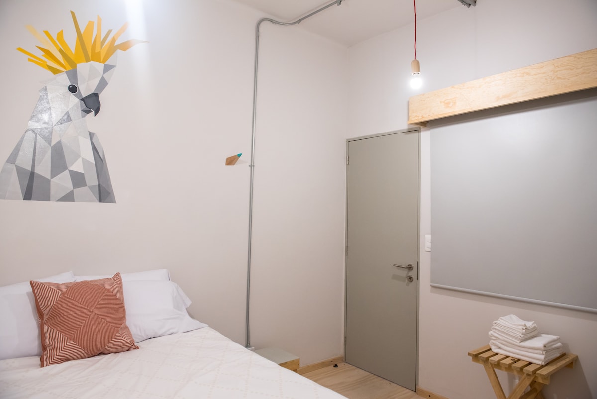 A bright room features a spacious bed adorned with white linens and a textured orange pillow. A playful geometric mural of a bird adds character to the wall. A simple, functional wooden shelf holds neatly folded towels, while a minimalist light fixture offers soft illumination.