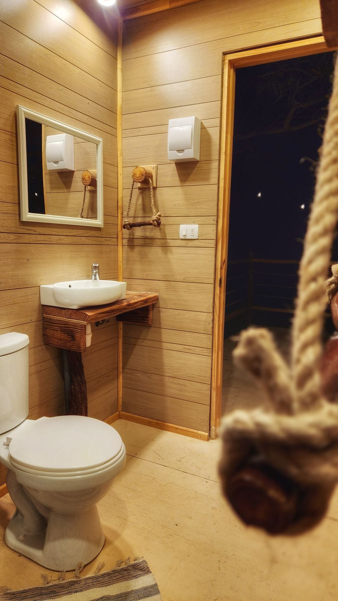 A well-appointed bathroom features natural wood accents and warm wooden walls. A modern sink is positioned above a handcrafted wooden vanity, and a clean toilet is visible. Soft lighting creates a cozy ambiance, while a mirror adds functionality to the space.
