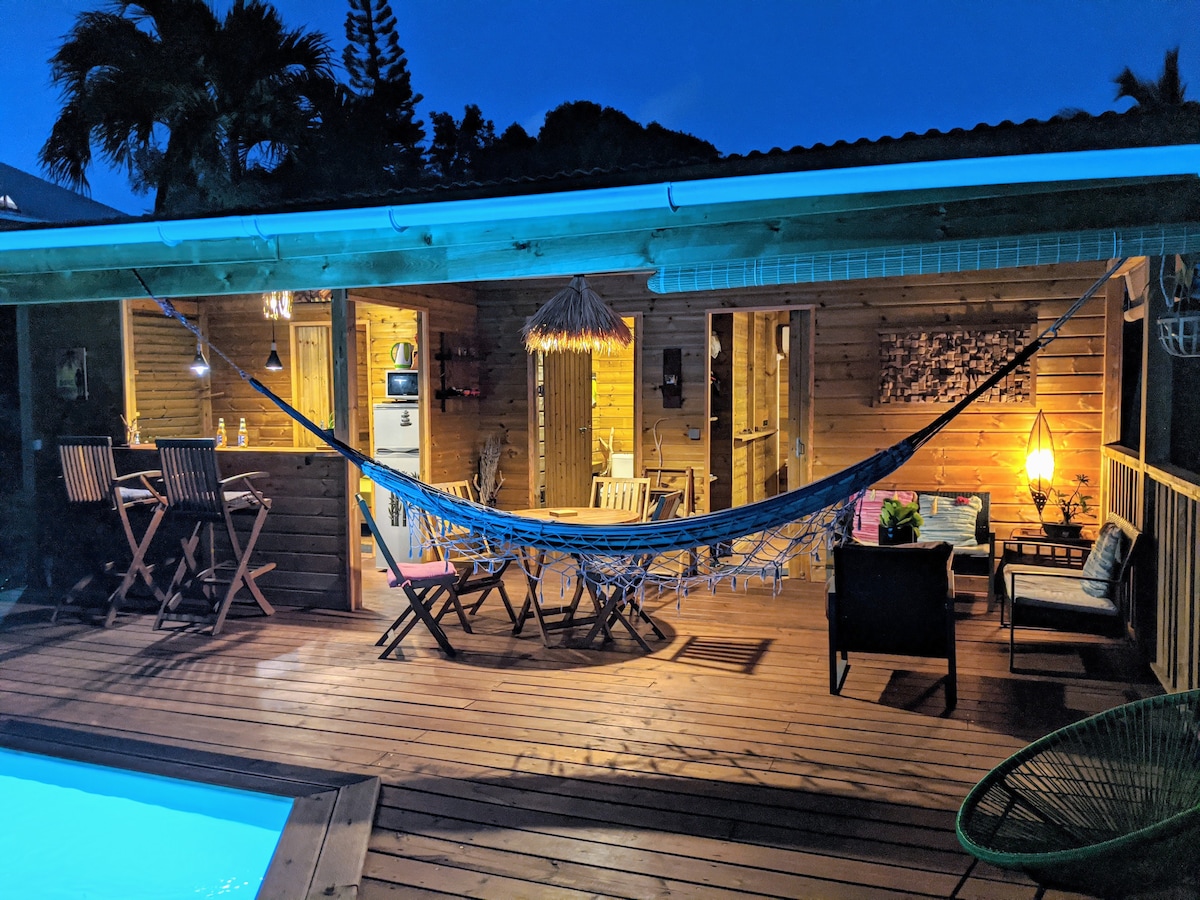An outdoor seating area is positioned by the private pool, featuring a hammock strung between two support posts. Wooden deck flooring is complemented by tropical plants in the background. The space is warmly lit by hanging lights, creating a relaxed atmosphere for evening gatherings.