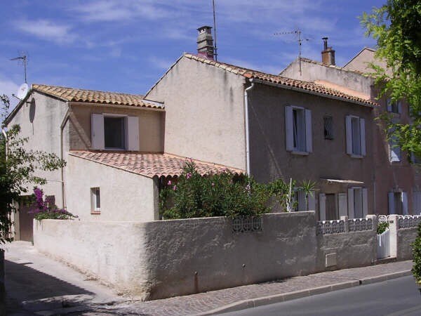 A charming village house is featured, showcasing a light-colored exterior with a tiled roof. Windows with shutters punctuate the walls, while flowering plants are visible in the garden area. The surrounding street is quiet, enhancing the village atmosphere.