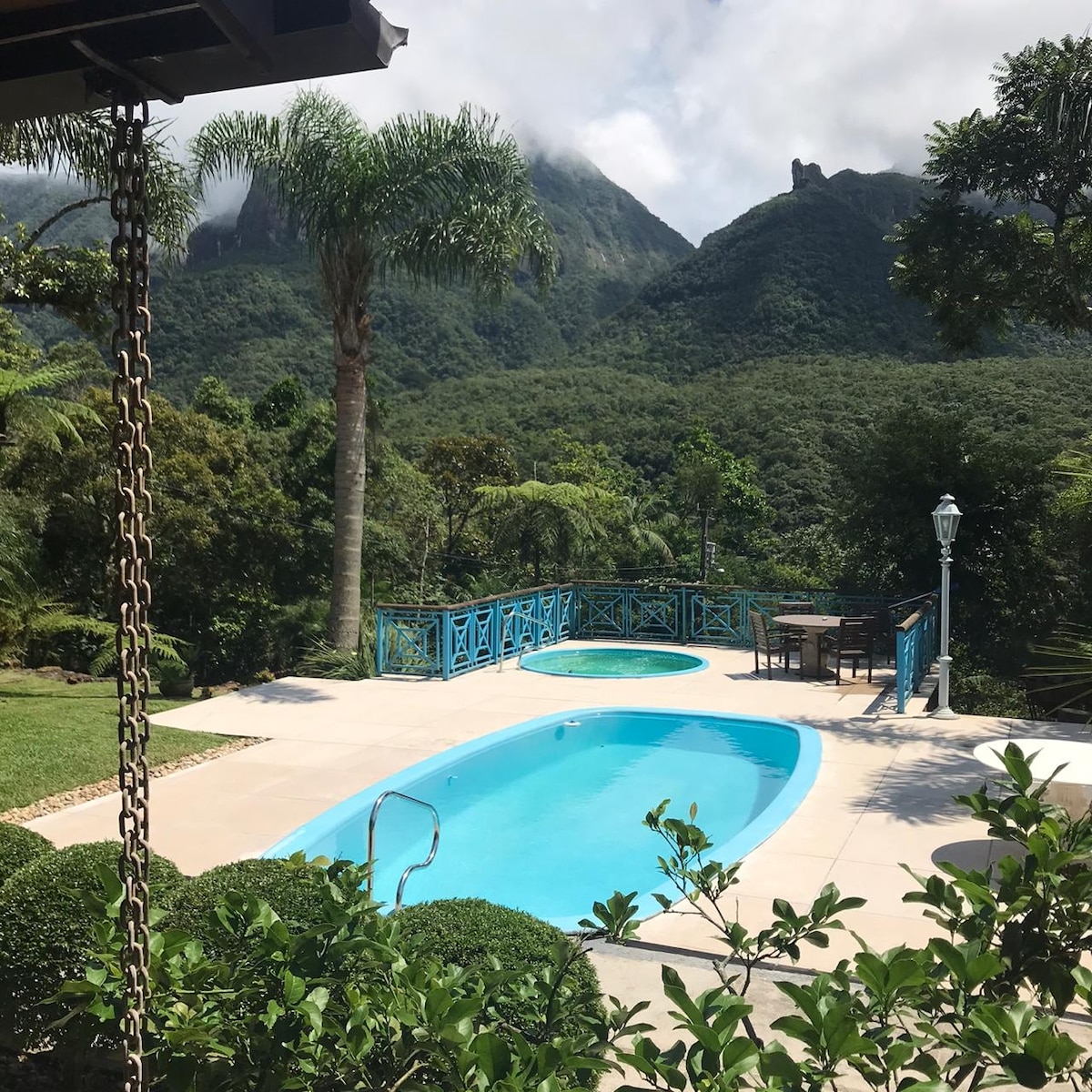 A tranquil outdoor area features a clear swimming pool surrounded by lush greenery. The backdrop showcases majestic mountains beneath a partly cloudy sky. Patio furniture is arranged nearby, providing a space for relaxation and enjoying the scenic view.