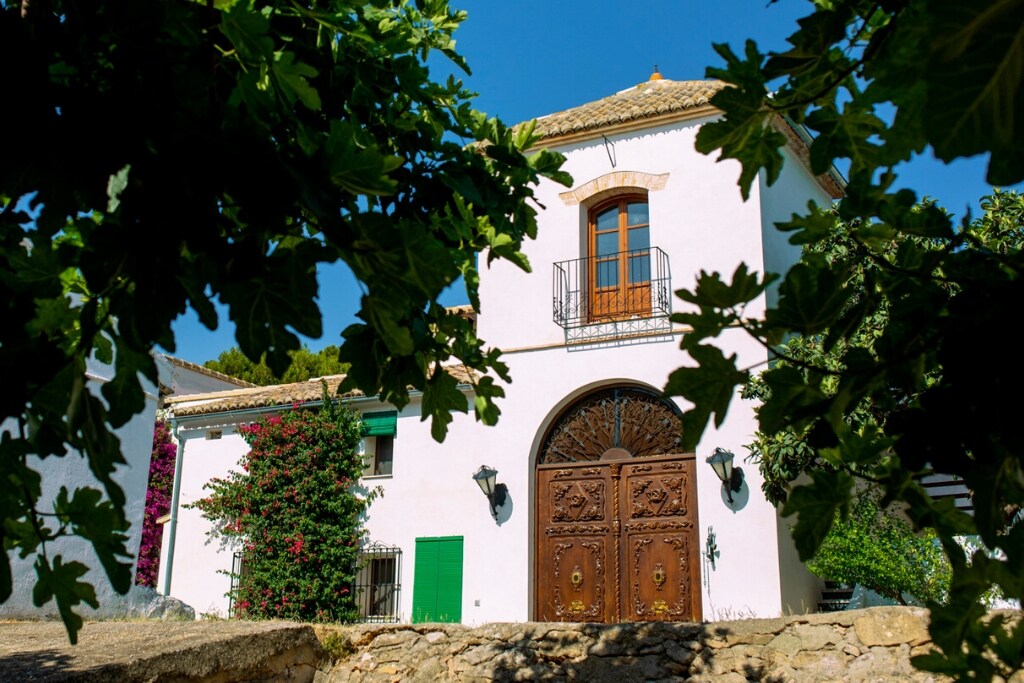 A charming farmhouse exterior is framed by lush greenery and vibrant bougainvillea. The well-crafted wooden doors feature intricate carvings, while a balcony above adds character. Sunlight highlights the cream-colored walls, creating an inviting entrance to the property.