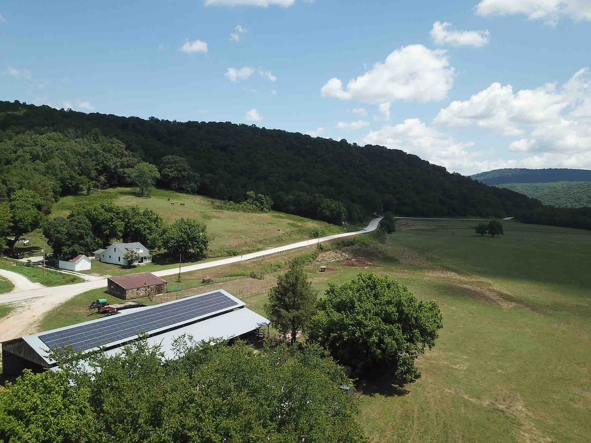 Aerial view captures the Villines' Farmhouse nestled against a backdrop of rolling hills and greenery. Solar panels atop a building reflect sunlight, while a winding road leads through expansive grassy fields. Trees offer a natural border, enhancing the serene rural landscape.