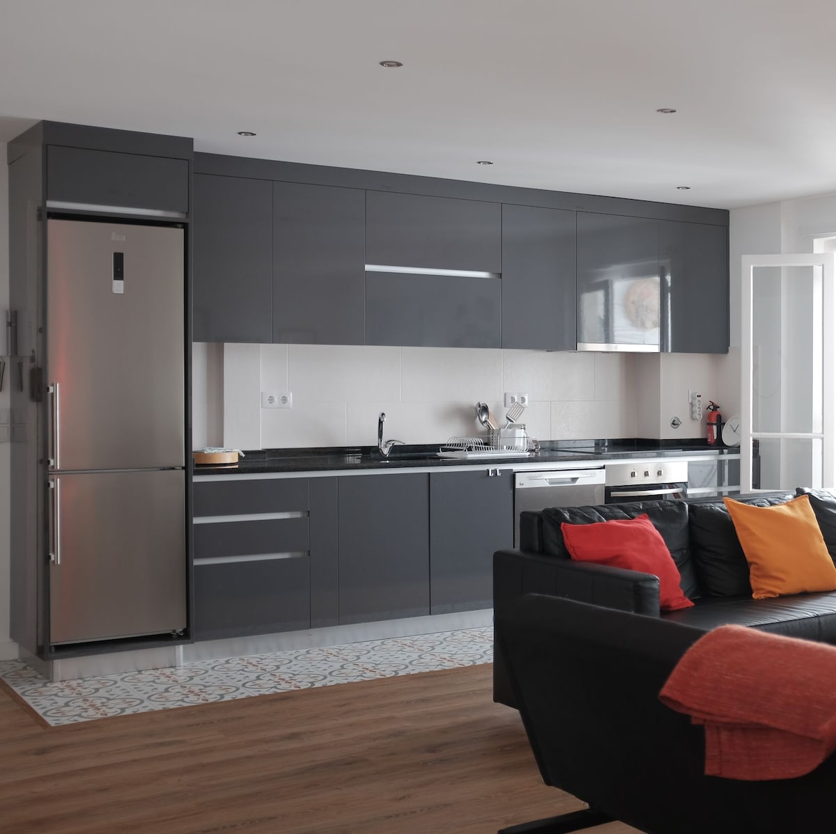 A modern kitchenette is equipped with sleek dark cabinetry and stainless steel appliances, including a refrigerator and dishwasher. A black sofa is positioned in front, adorned with colorful cushions. Natural light streams in through a window, enhancing the space's contemporary design.