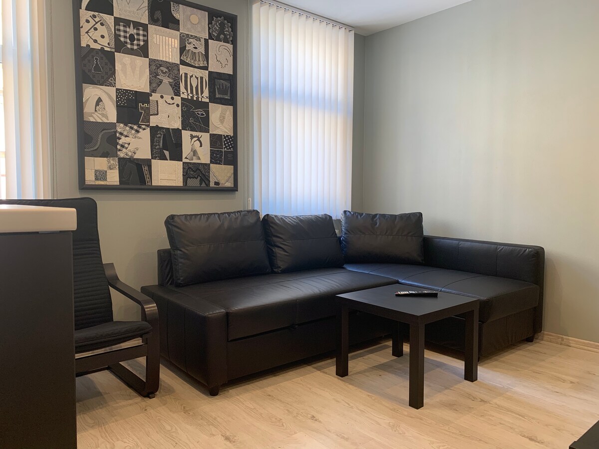 A cozy living area features a dark leather sectional sofa adjacent to a minimalist black coffee table. Natural light filters through vertically striped window coverings, illuminating the space. A patterned wall art piece adds a modern touch to the room.