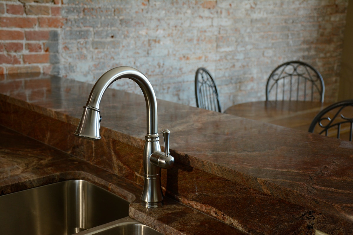 A modern kitchen features a polished granite countertop with an elegant stainless steel faucet. Exposed brick walls provide a rustic backdrop, while two black metal chairs are positioned at a nearby wooden dining table, enhancing the inviting atmosphere of this remodeled loft.