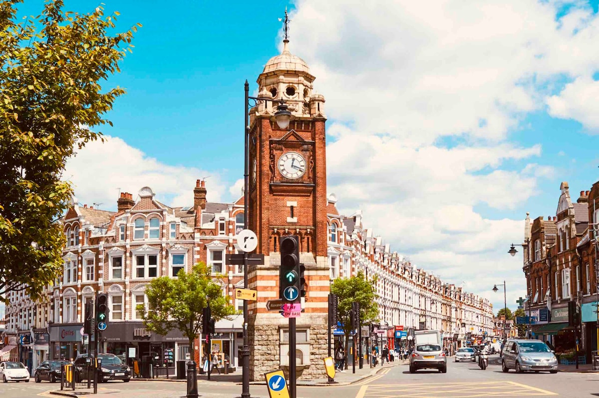 A historical clock tower stands prominently at the intersection, surrounded by a row of shops and cafes. Lush green trees line the street, with vehicles passing by under a bright blue sky adorned with scattered clouds.
