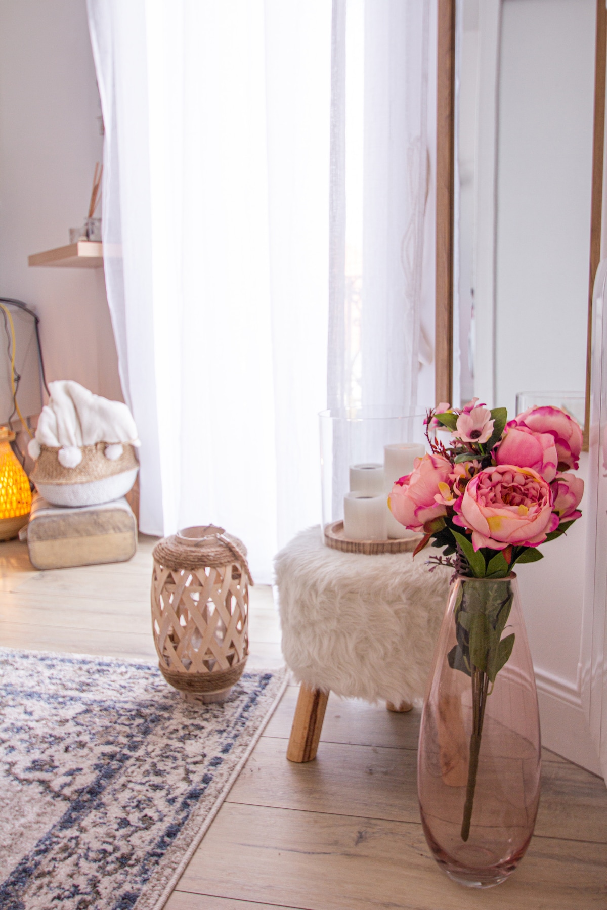 A bright, airy corner displays a large vase filled with pink peonies next to a soft, plush stool. Nearby, a candle centerpiece is placed atop a furry surface. Natural light filters through sheer curtains, creating a warm and inviting space.