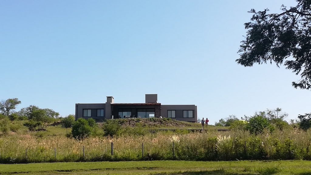 A modern house is situated on a hillside, surrounded by tall grasses and greenery. Large windows offer expansive views, while the structure features a minimalist design. The clear blue sky enhances the outdoor setting, indicating a peaceful, rural environment.