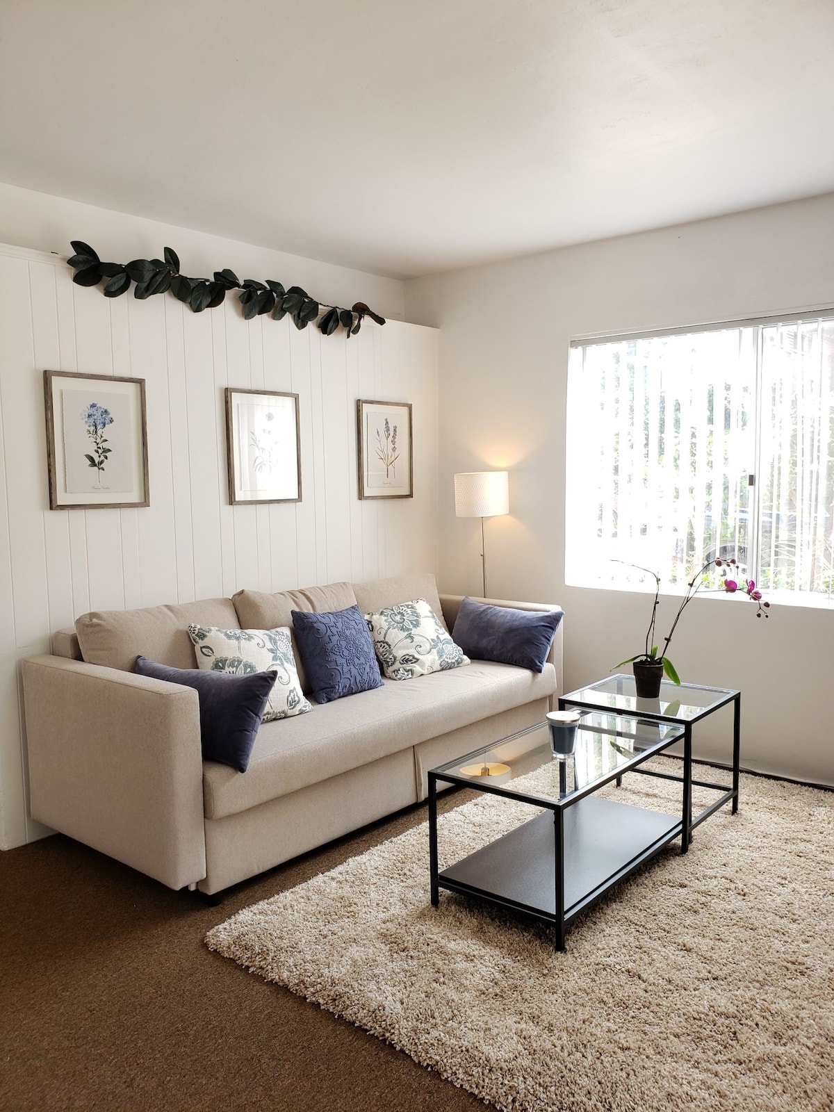 A cozy living area features a light-colored sofa adorned with patterned cushions, complemented by a rectangular coffee table. Natural light enters through a window adorned with vertical blinds, while a small plant adds a touch of greenery to the space. Framed botanical artwork is displayed on the wall.