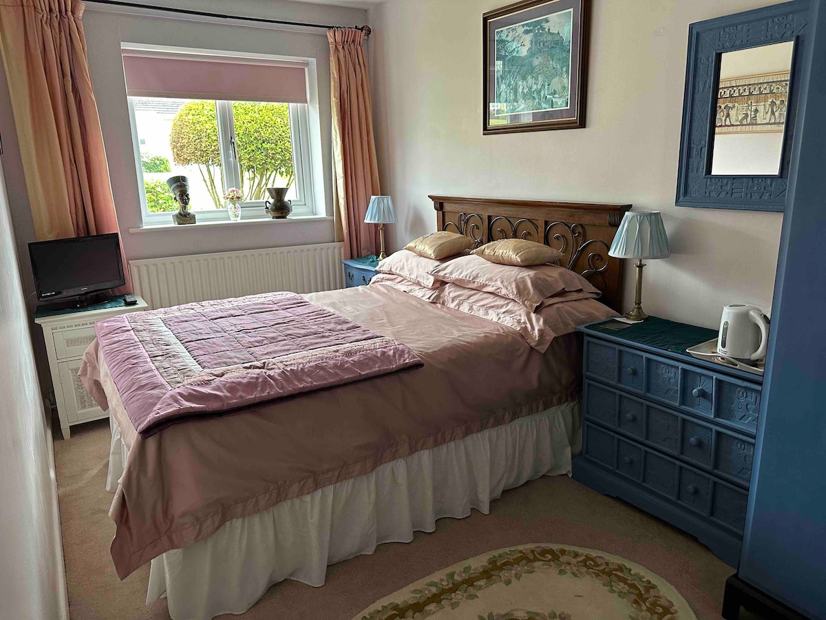 A bedroom features a comfortably made bed with a pink bedspread and multiple pillows. A small television is positioned on a dresser, and bedside lamps provide soft lighting. A window with light curtains and decorative items enhances the ambiance, creating a serene atmosphere.