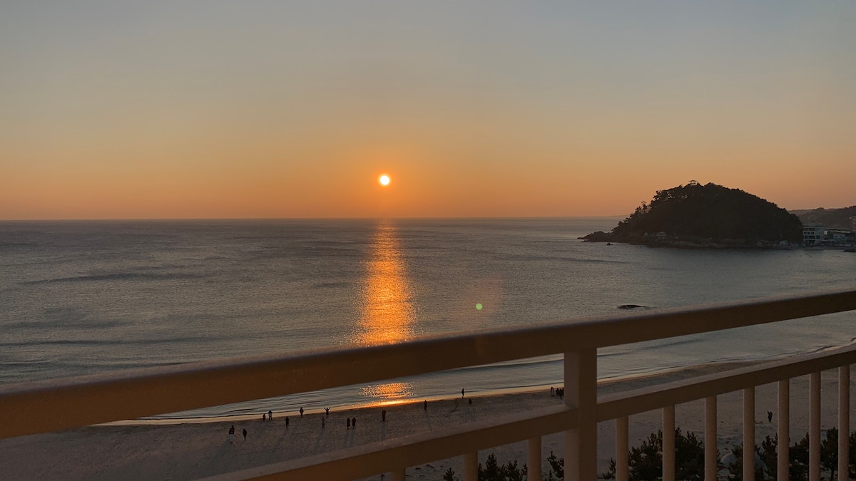 A serene sunset is captured over the calm ocean, with the sun reflecting on the water's surface. Silhouettes of people can be seen walking along the beach, framed by the gentle outline of a nearby island.