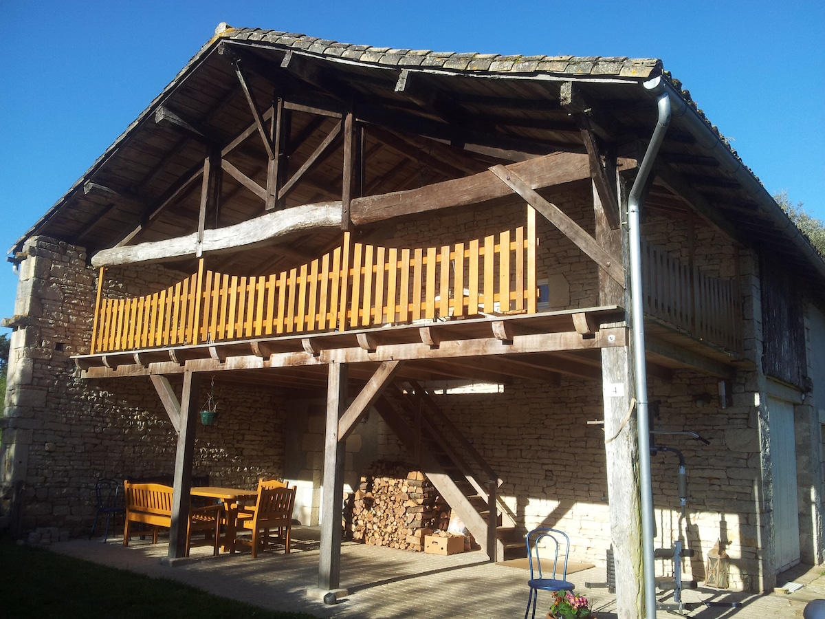 An outdoor view of a rustic house features a spacious wooden balcony supported by sturdy beams. Below, a seating area is accompanied by a table and stacked logs, with direct access to an exterior staircase leading up to the balcony.