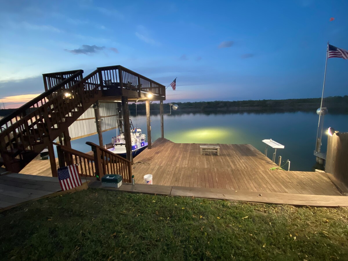 A spacious wooden dock is seen at twilight, featuring a well-lit area suitable for gatherings. A staircase leads to an elevated platform, while flags adorn the dock. The calm waters reflect the evening sky, enhancing the serene atmosphere of the Colorado River.