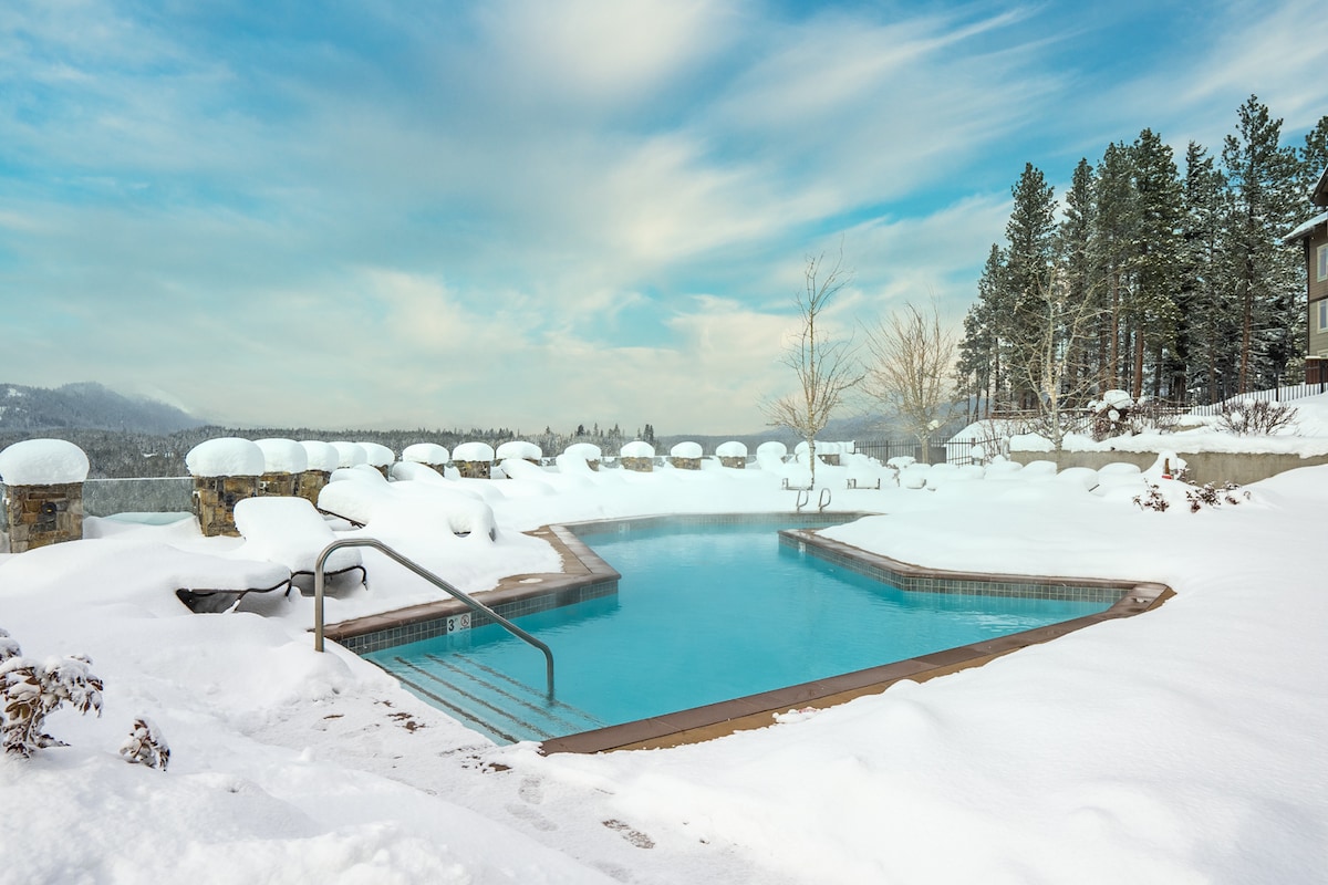 The outdoor pool is surrounded by a winter landscape, featuring snow-covered lounge chairs that complement the serene blue water. Snow blankets the ground, and trees in the background are dusted with white, creating a peaceful atmosphere.
