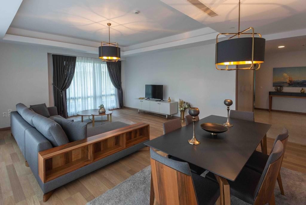 A modern living area features a gray sectional sofa and a stylish dining table with wooden accents. Large windows allow natural light to fill the space, while elegant light fixtures and decorative items enhance the contemporary decor. A television is positioned against the wall.