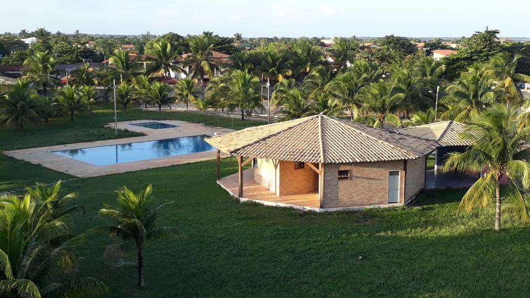 A spacious outdoor area is surrounded by lush greenery. A pool occupies one side, while a covered structure is positioned nearby, featuring a thatched roof. Palm trees are scattered throughout, providing shade and a relaxed atmosphere.