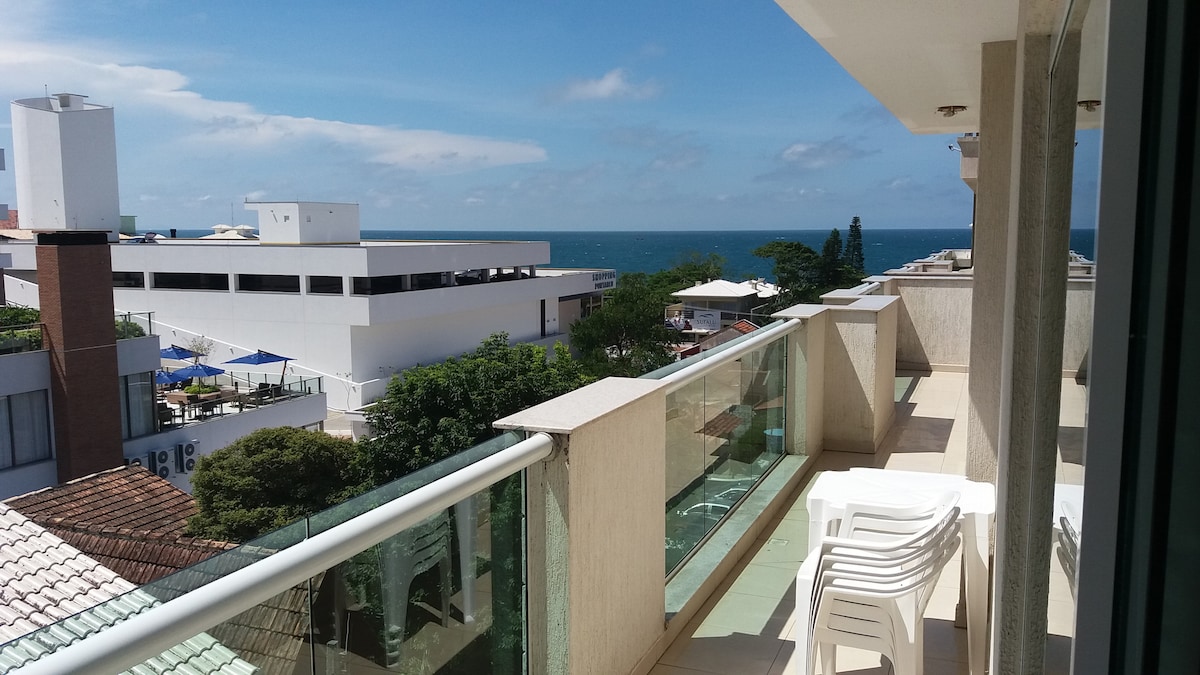 A spacious balcony with a glass railing overlooks the ocean, allowing for a clear view of the sea. White plastic chairs are positioned on the balcony, inviting relaxation. Surrounding greenery and nearby buildings are visible, contributing to the serene atmosphere.