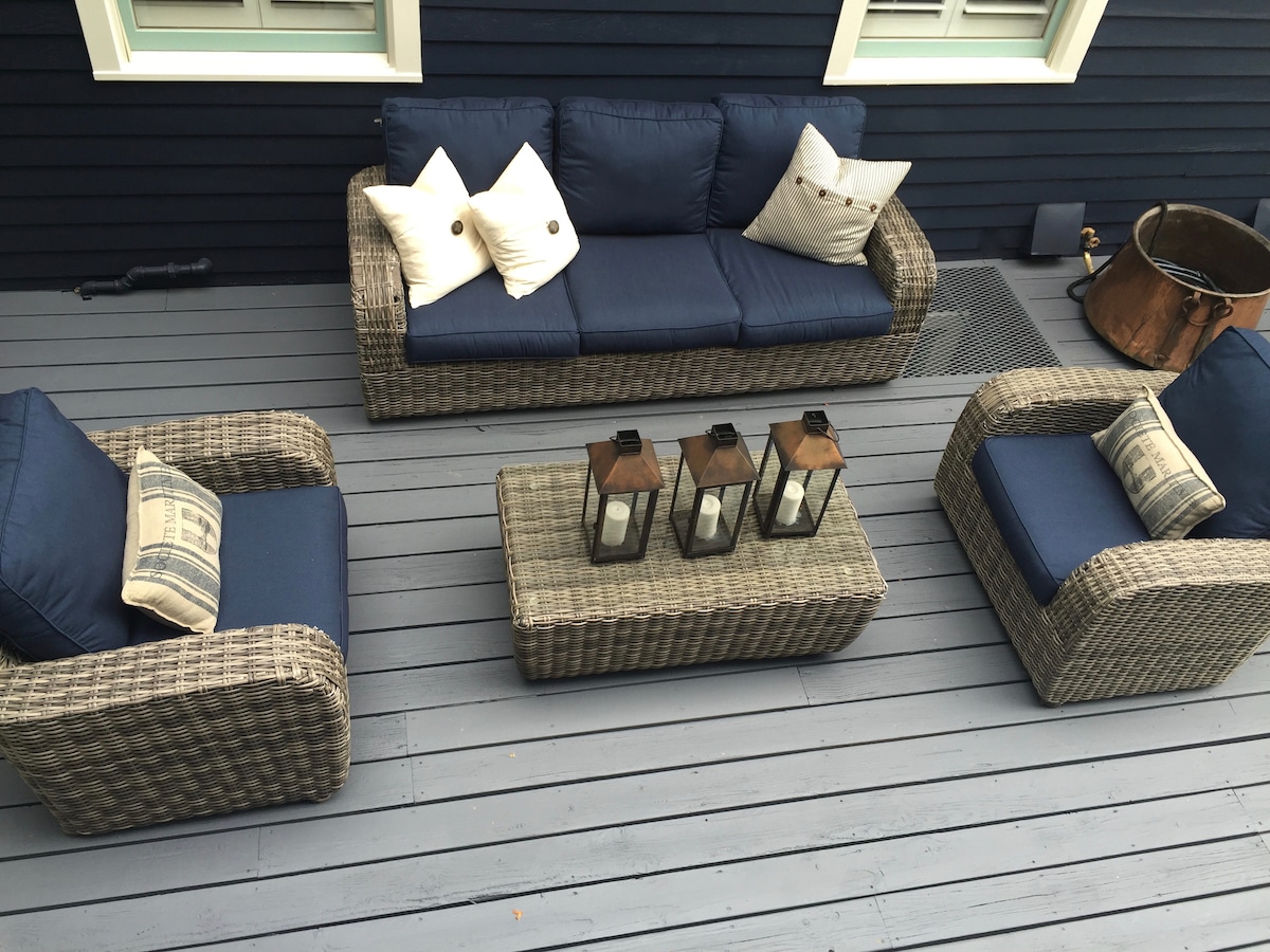 An outdoor seating area is presented with a woven sectional sofa and two matching armchairs, all featuring navy blue cushions. A rectangular coffee table holds three lanterns, adding a decorative touch. The space is set on a gray deck, surrounded by a well-maintained exterior.