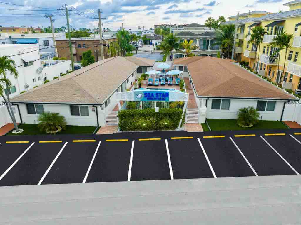 Aerial view of a well-kept property featuring two single-story buildings surrounded by palm trees and vibrant landscaping. The central area includes a pool with chaise lounges and dining tables. A clear parking area with marked spaces is visible, enhancing accessibility.