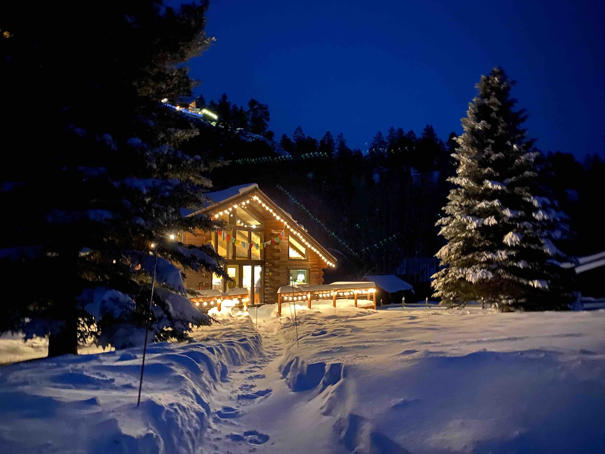 The cabin is nestled among snow-covered trees, illuminated by warm lights along the roofline. A clear path through the snow leads towards the front entrance, highlighting the cabin’s welcoming presence in a serene winter landscape. The dark sky enhances the cozy ambiance.