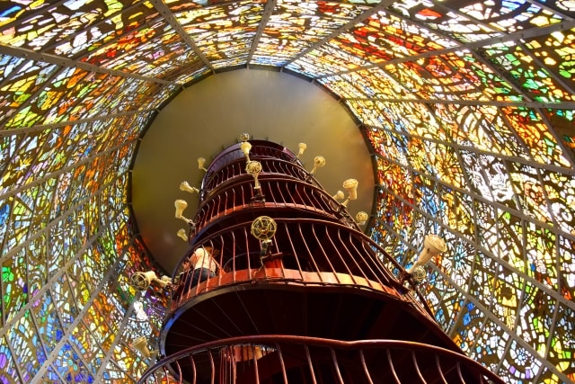 A spiraling staircase ascends through a vibrant dome filled with intricate stained glass. The interplay of colorful light casts dynamic patterns on the staircase, enhancing the surrounding atmosphere. Decorative railings and light fixtures are visible along the ascent.