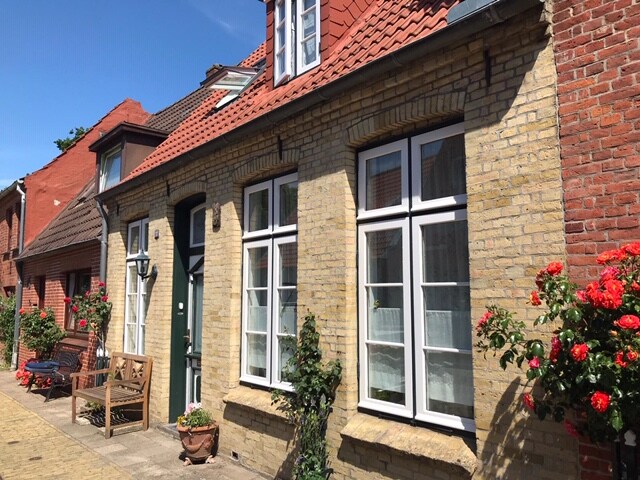 A charming brick facade displays multiple large windows with white frames, allowing natural light to flood the interior. Flower pots with vibrant roses adorn the entrance, enhancing the inviting atmosphere of this quaint, historic property.