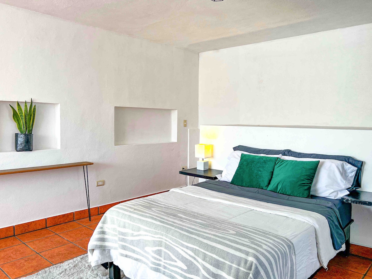 A bright sleeping area is characterized by neutral walls and terracotta tile flooring. A spacious bed is dressed in crisp white linens with green accent pillows. A small side table features a lamp, and a potted plant adds a touch of greenery.