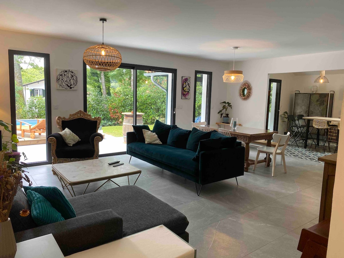 A spacious living area is highlighted by large glass doors leading to a garden. Comfortable seating arrangements include a dark sofa and a striking armchair. Natural light fills the space, complemented by modern decor and a dining area featuring a table with chairs.