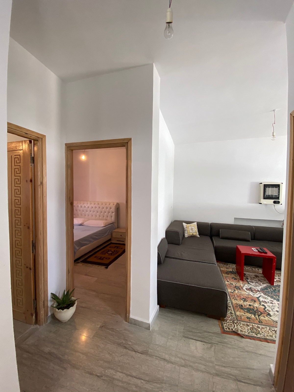 A spacious living area is seen featuring a comfortable gray sofa arranged around a small red table. Light flows in from a nearby window, illuminating the tiled floor. Doorways lead to two bedrooms, enhancing the apartment's layout.