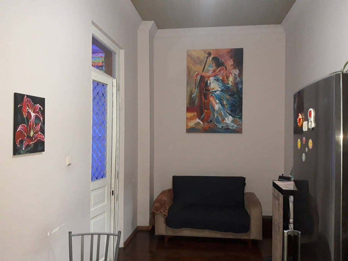 The living area features a cozy sofa, positioned against a light-colored wall, with a vibrant painting above. A stainless steel refrigerator and a small black table are present, contributing to a functional space. Art pieces adorn the walls, adding character to the room.