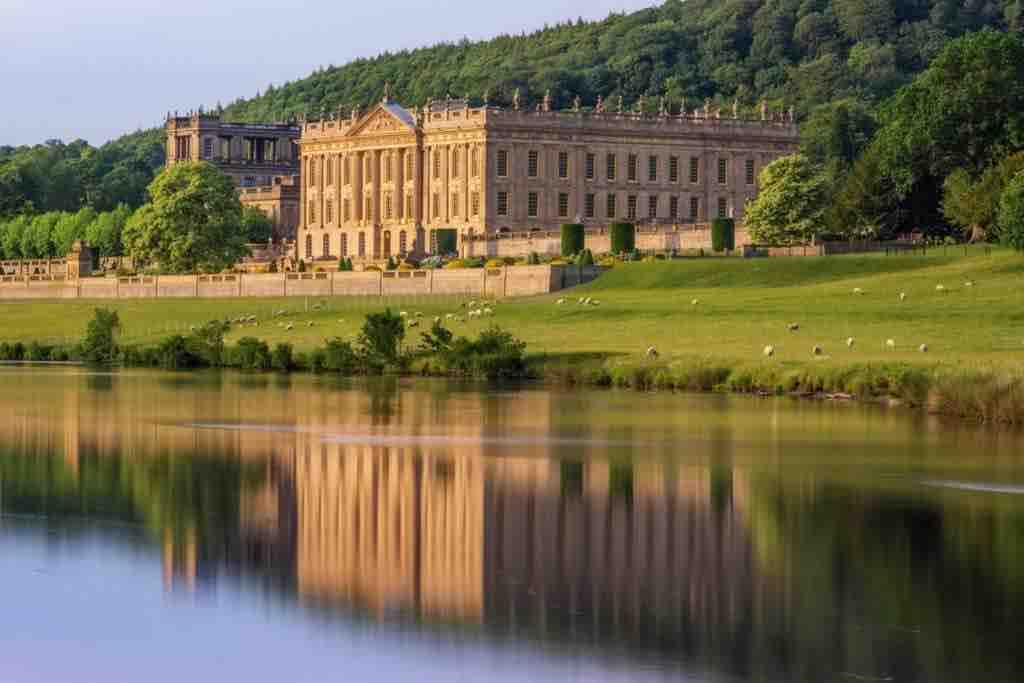 A grand historical house is depicted beside a serene river, surrounded by lush greenery and rolling hills. The building's facade reflects in the water, emphasizing its architectural details and the surrounding natural beauty. Sheep can be seen grazing in the fields nearby.