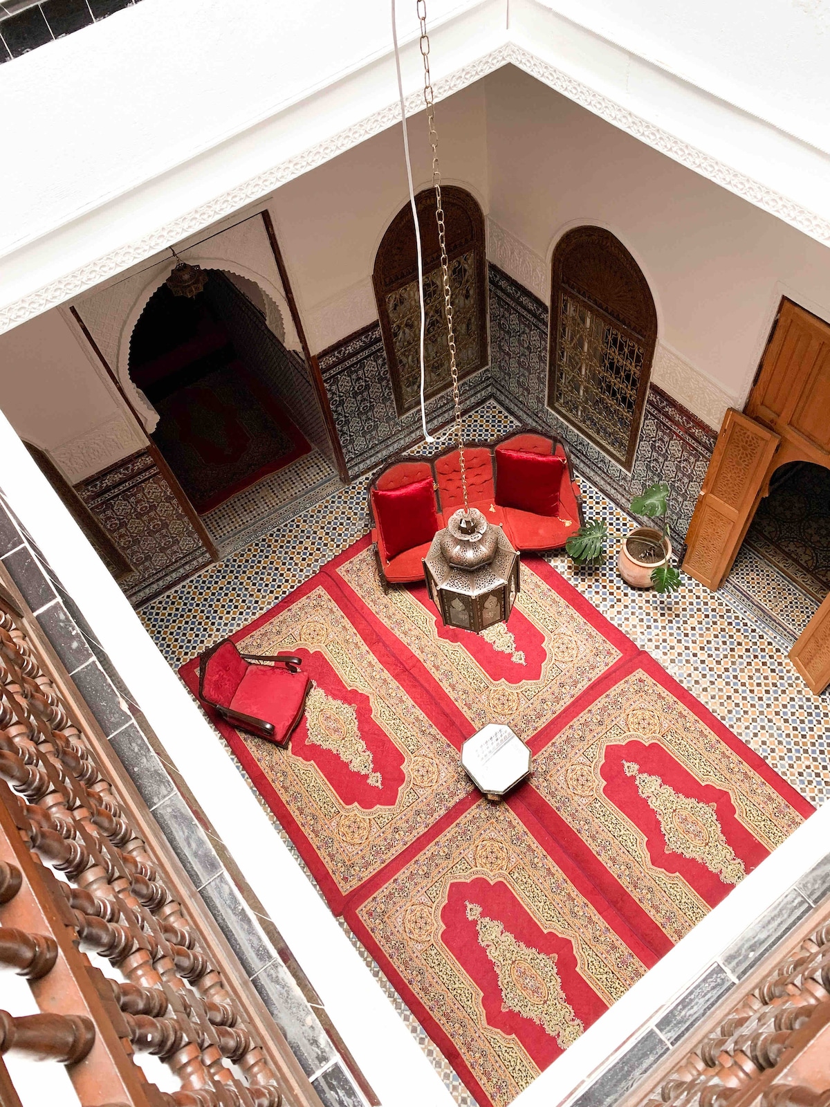 The central patio features intricate tile work and traditional decor, with two red chairs positioned alongside a small table. A decorative lantern hangs from the ceiling, complemented by a green plant. The surrounding walls are adorned with patterned tiles, enhancing the space's character.