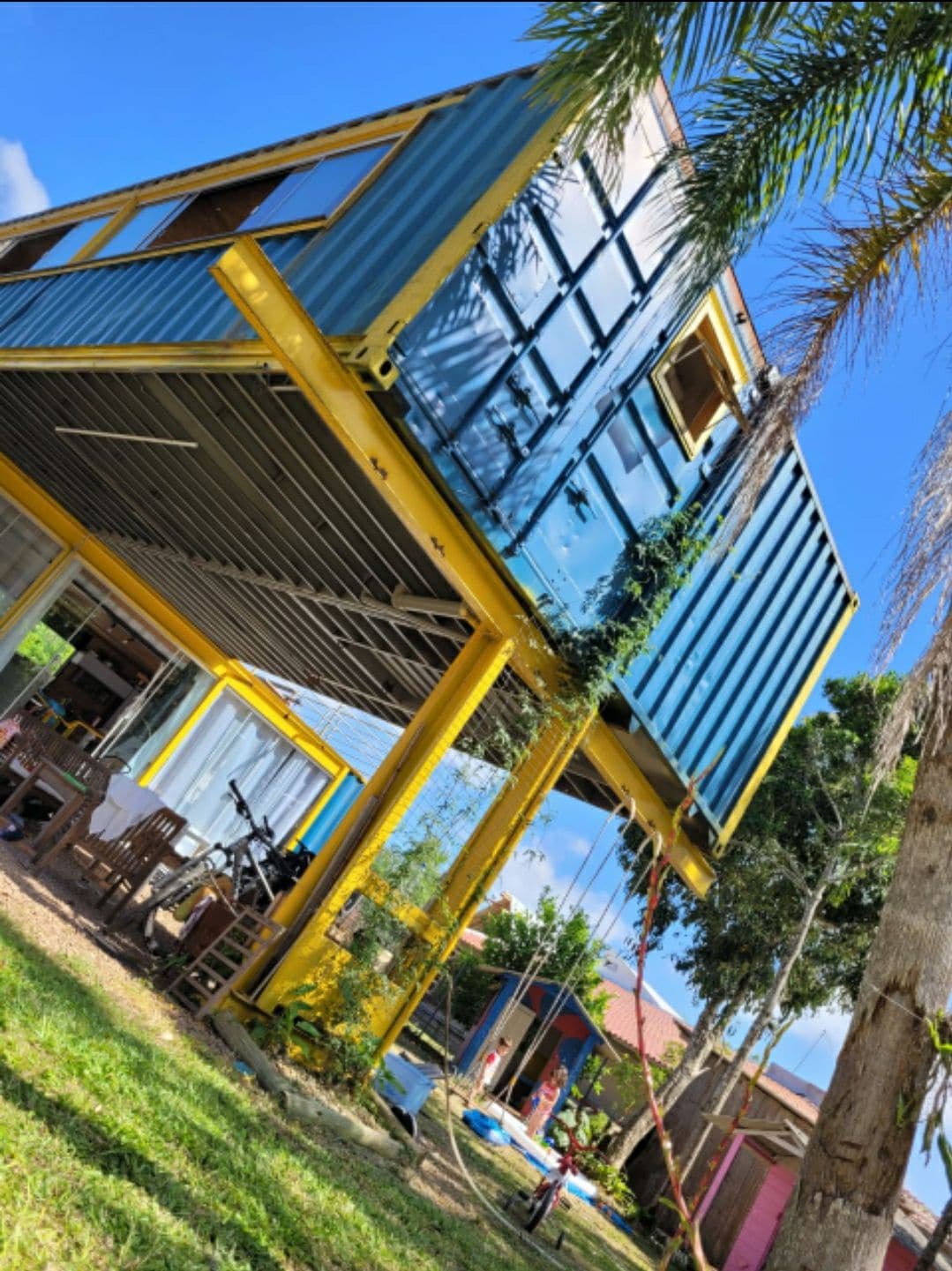 A vibrant blue shipping container is elevated and supported by yellow pillars, surrounded by lush greenery. The structure features large windows and an outdoor seating area, with glimpses of bicycles and play areas in the backyard.