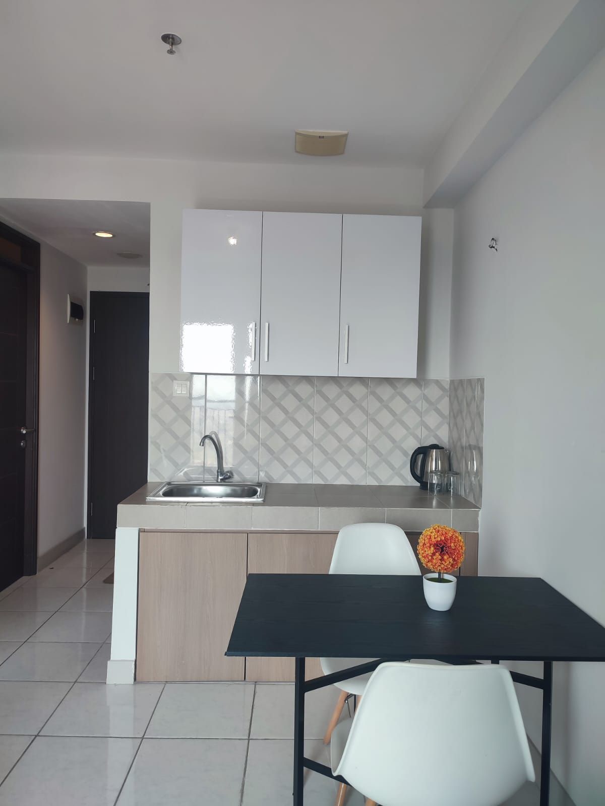 A compact kitchen area features light-colored cabinetry and a tiled backsplash. A stainless steel sink and kettle are included. A small dining table with two white chairs is set for meals, adorned with a vibrant flower centerpiece.