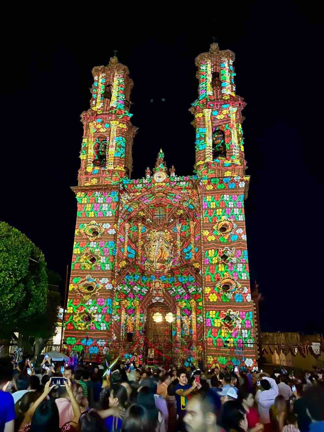 A illuminated church facade, adorned with vibrant light projections, displays intricate floral patterns and religious figures. The surrounding area is filled with a gathering of people, experiencing the festive atmosphere at night, with trees and a clear sky visible in the background.