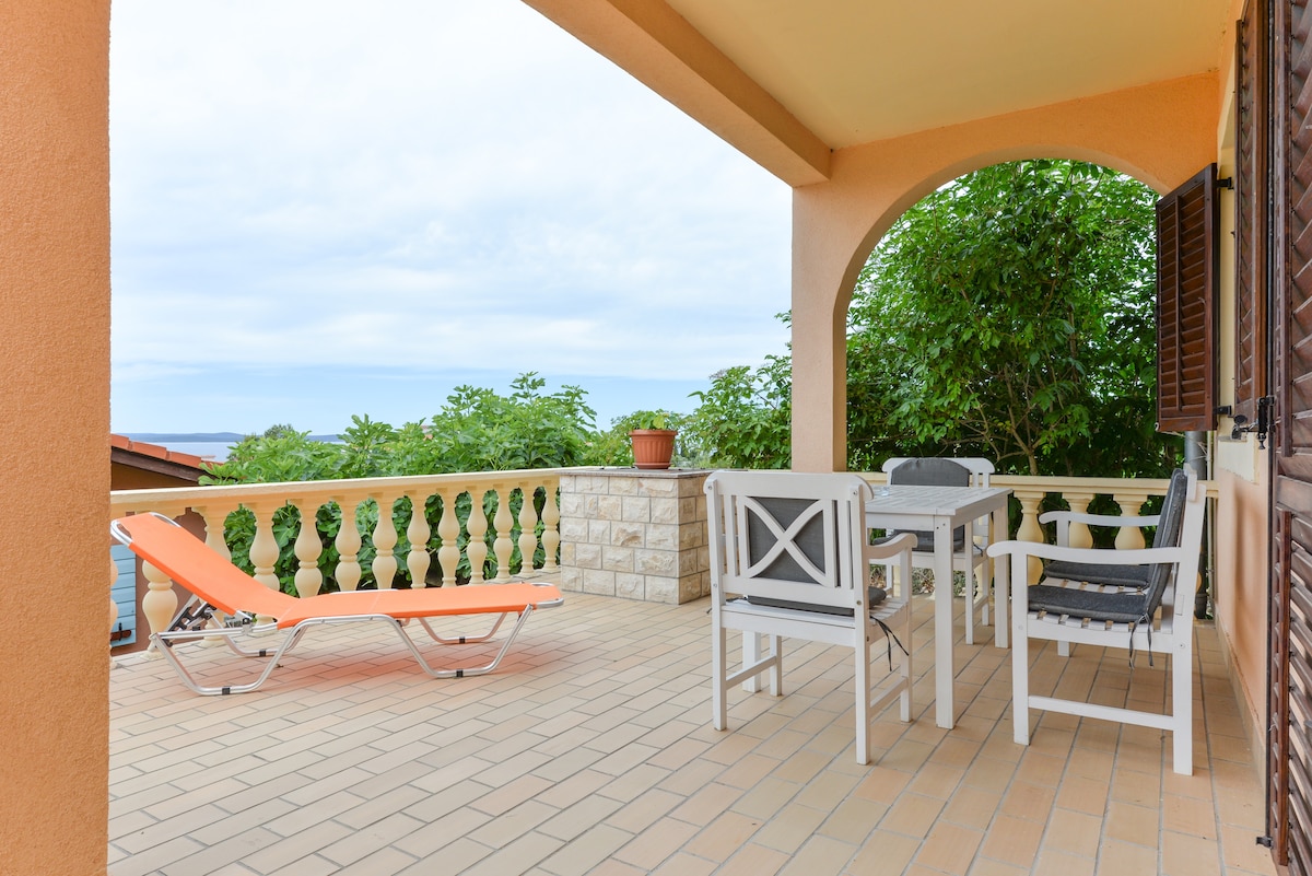 A spacious terrace is featured, furnished with a dining table and chairs, alongside a lounge chair. Lush green foliage is visible in the background, adding to the serene environment. A distant sea view can be seen, framed by the terrace's elegant archways.