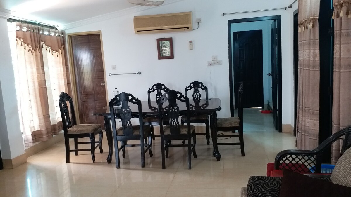A dining area is featured with a dark wooden table surrounded by six intricately designed chairs. Natural light enters through two large windows, which are dressed with light curtains. A door leads to another room, complementing the spacious layout.